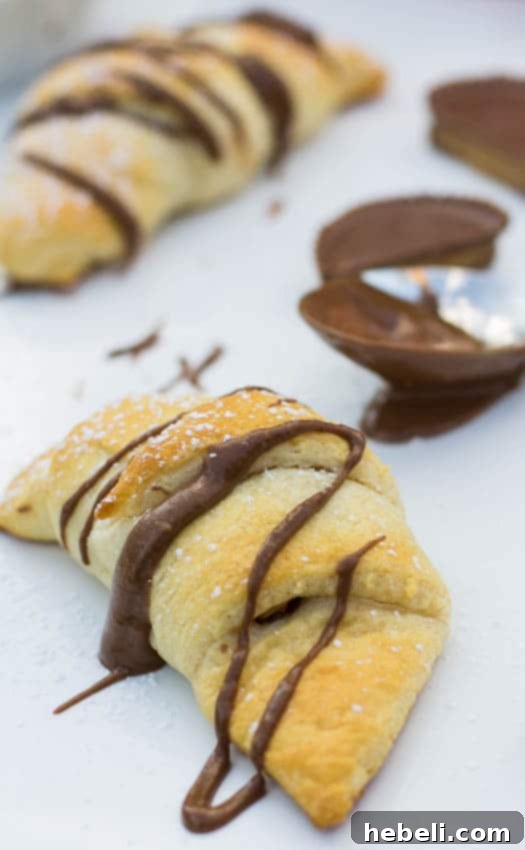 Sweet & Salty Reese's Crescent Rolls 3 A close-up of a warm, golden crescent roll, split open slightly to reveal the melted Reese's peanut butter cup inside, generously drizzled with chocolate.