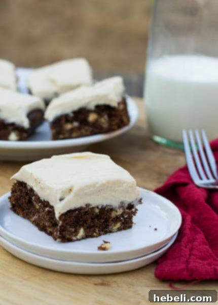 A close-up shot of a single gingerbread brownie adorned with a generous swirl of cream cheese frosting on a white plate, highlighting its rich texture and festive appeal, with more brownies visible in the blurred background.
