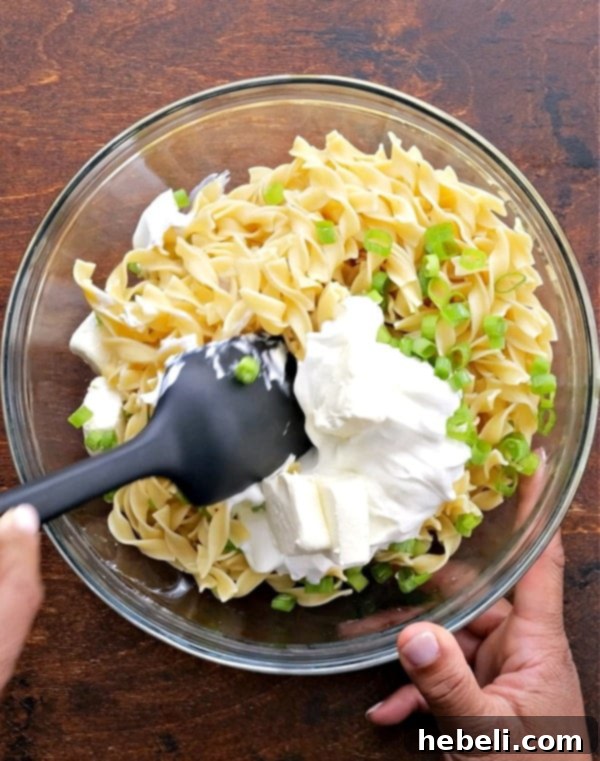 Comforting Ground Beef Supper 6 A bowl filled with cooked egg noodles, sour cream, cream cheese, and green onions, being mixed to create the creamy noodle layer.