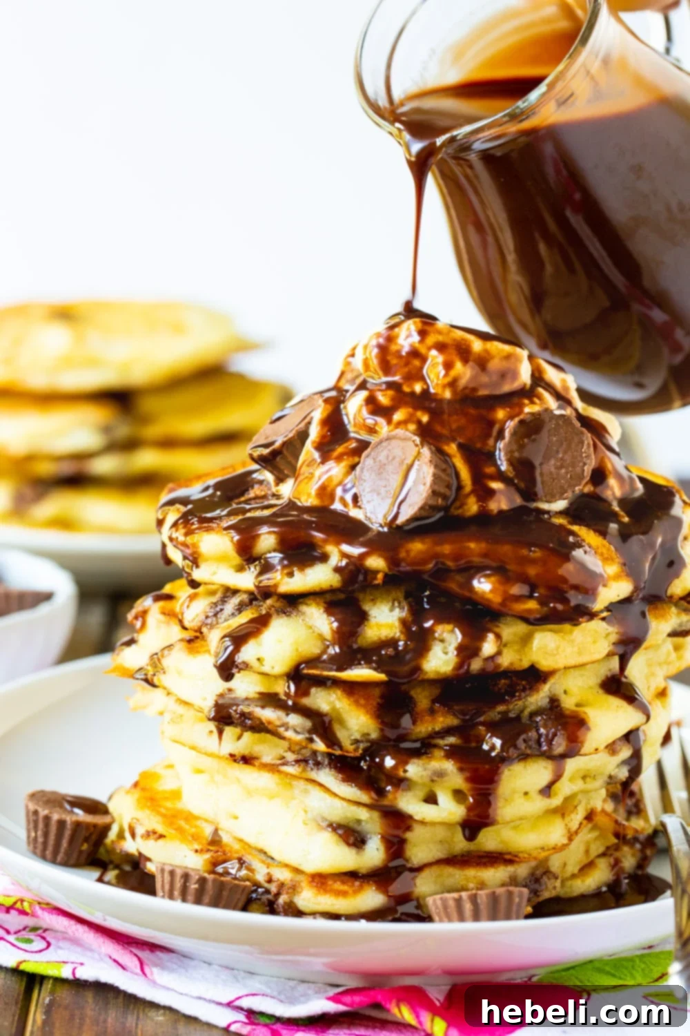 Homemade chocolate sauce being artfully poured over a beautifully arranged stack of Peanut Butter Cup Pancakes, garnished with whipped cream.