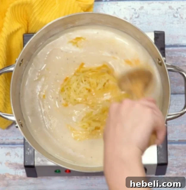 Golden Baked Ham Macaroni 5 Image showing shredded cheese being added to the thick, bubbling béchamel sauce in a pot, ready to melt.
