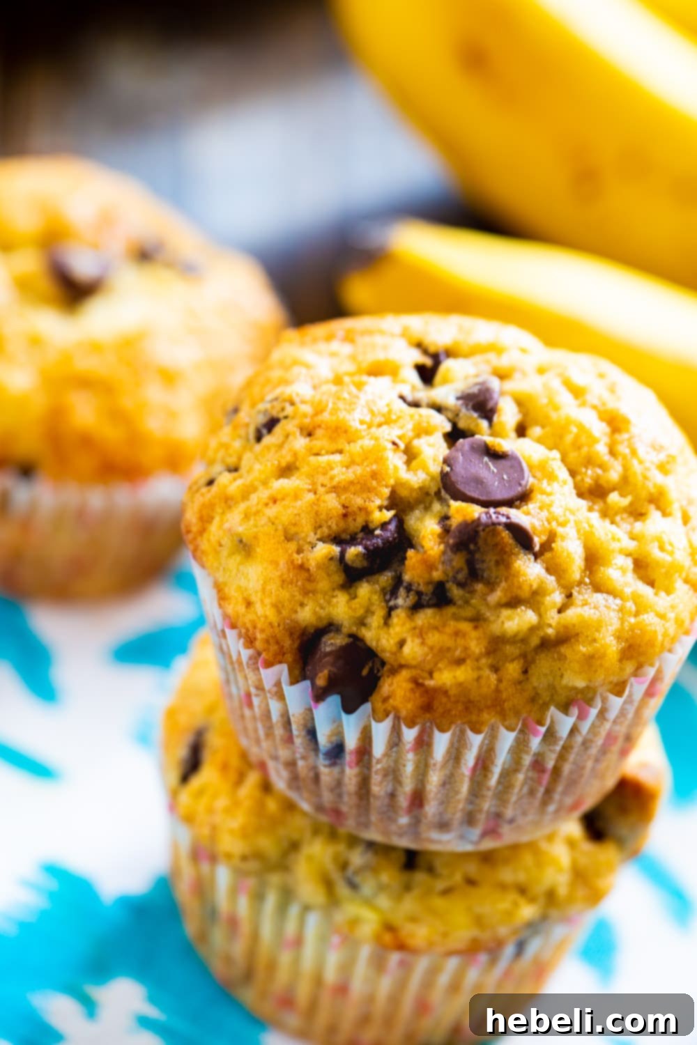 Two moist banana chocolate chip muffins stacked, with fresh ripe bananas in the blurred background, hinting at the key ingredient.