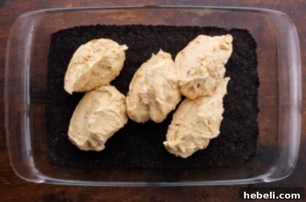 Spreading the thick peanut butter and cream cheese layer, speckled with Butterfinger crumbles, over the Oreo cookie crust in a baking dish.
