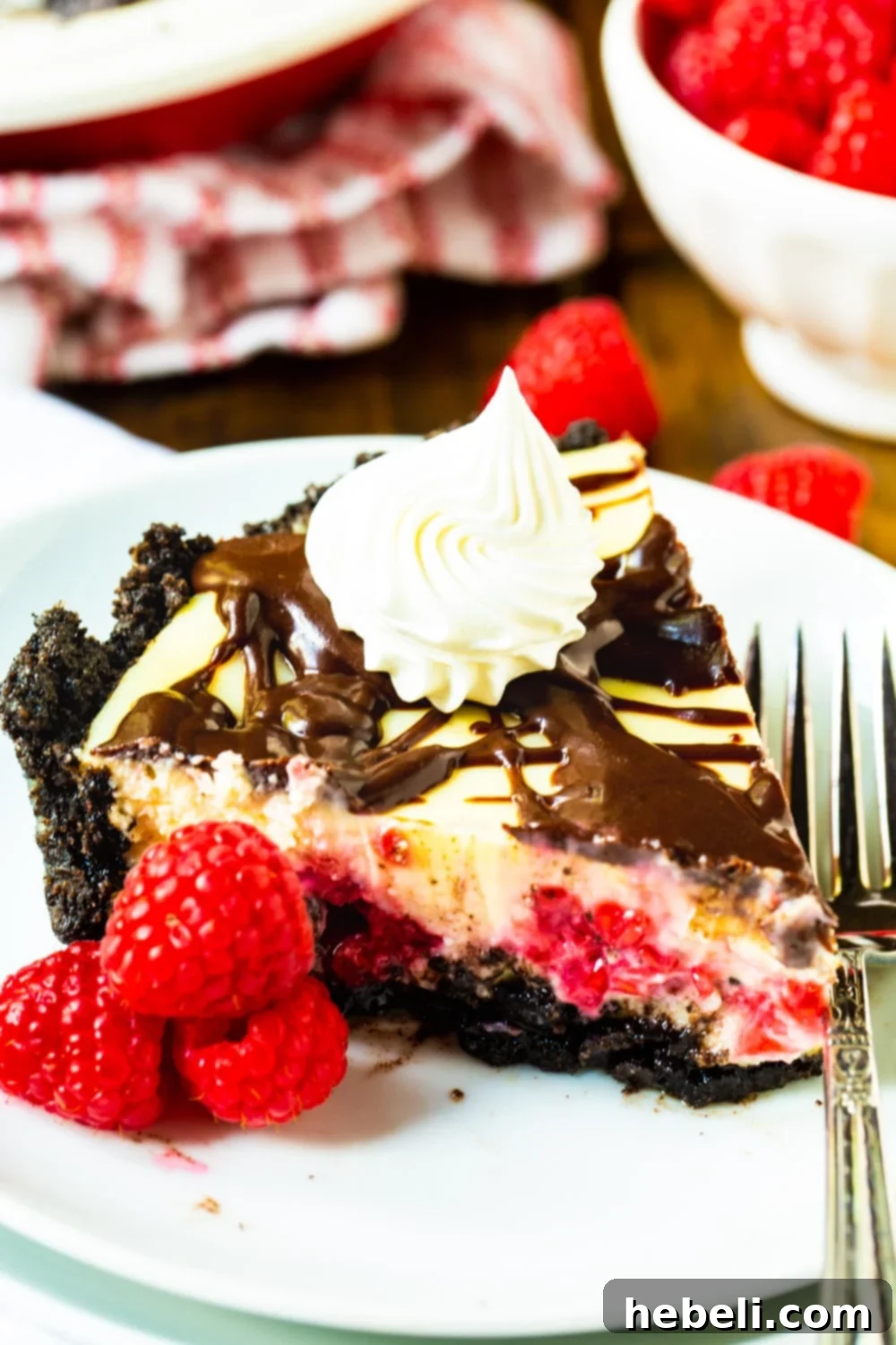 A close-up of a slice of Raspberry Cream Cheese Pie, showing the distinct layers of Oreo crust, creamy filling, and fresh raspberries, drizzled with chocolate glaze.