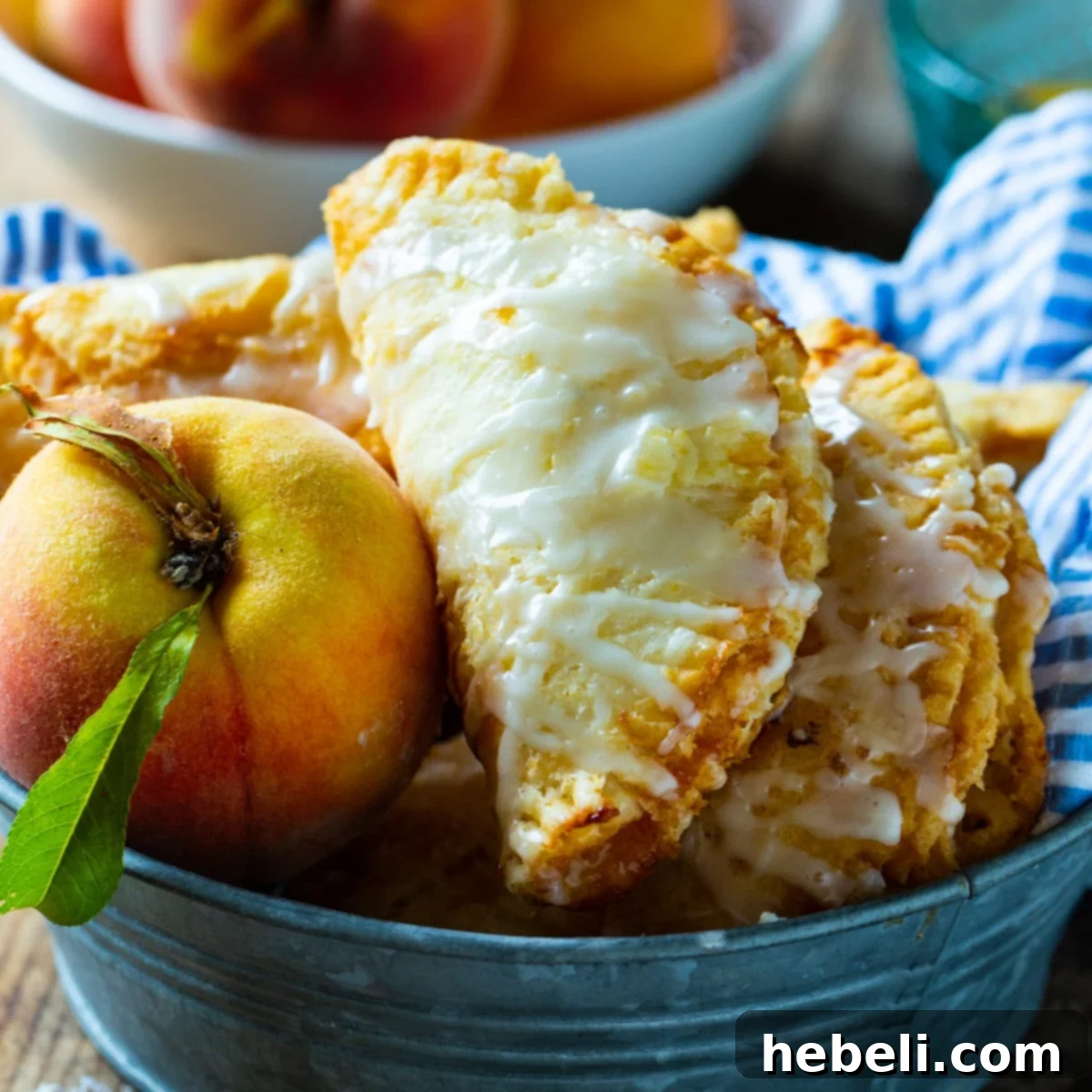 Golden Crispy Peach Turnovers 2 Golden brown Fried Peach Pies arranged beautifully in a rustic metal tin, with a fresh peach placed beside them, evoking a sense of sweet summer indulgence.