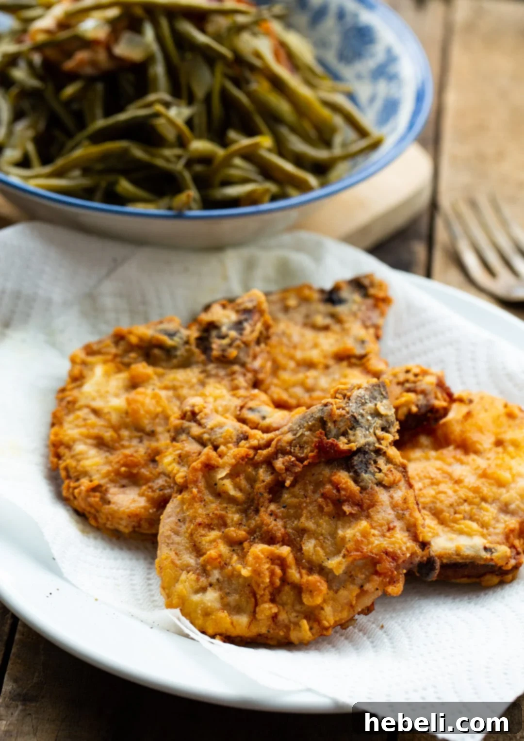 A plate of two perfectly fried Southern Fried Pork Chops, served alongside a bowl of vibrant green beans, showcasing a classic Southern meal.