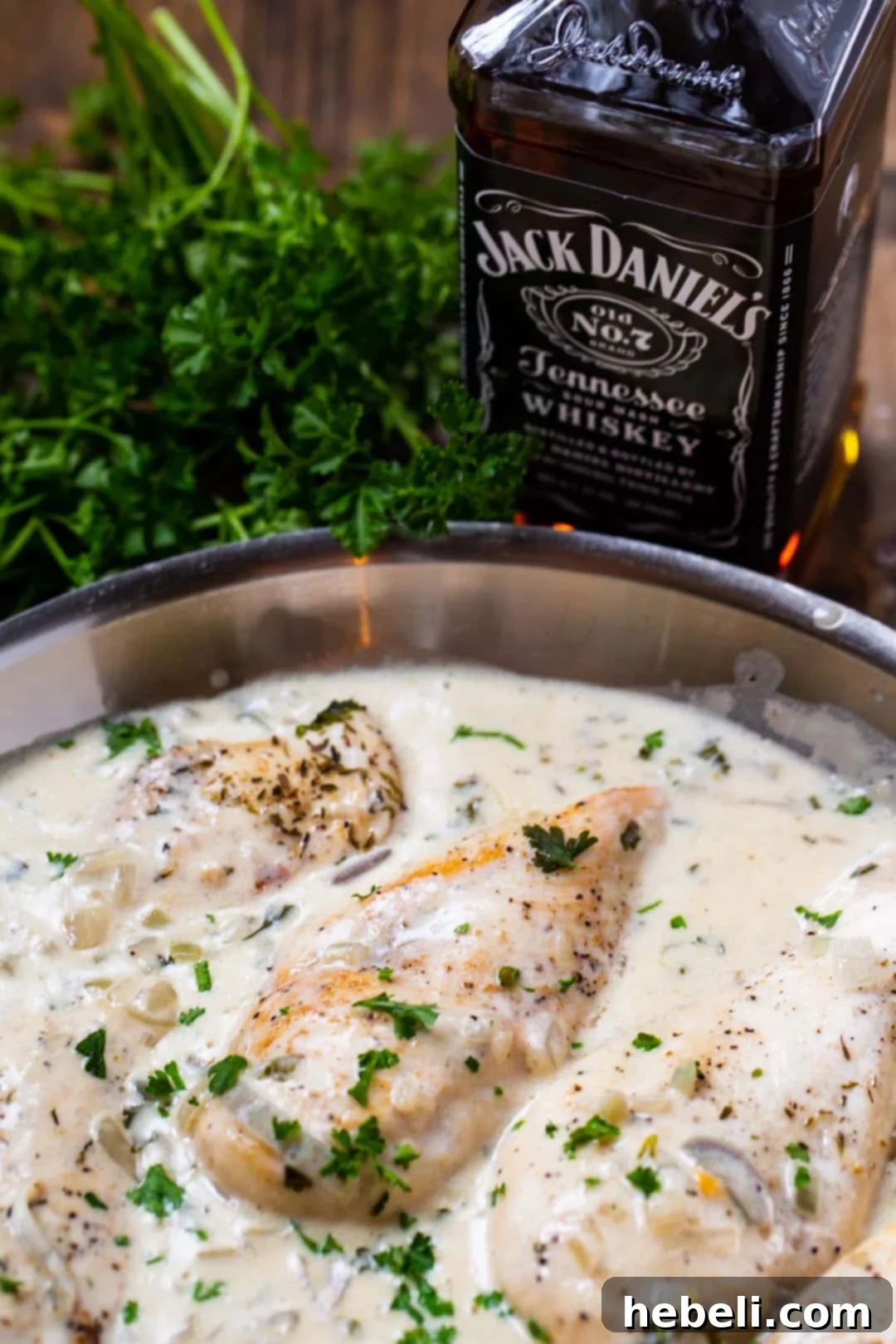 Close-up of golden-brown chicken and creamy sauce in a skillet, with a bottle of Jack Daniels whiskey in the background, hinting at the key ingredient.