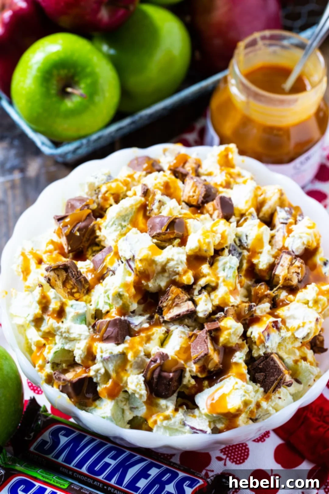 Close-up of Snickers Caramel Apple Salad in a bowl, surrounded by whole apples and full-sized Snickers bars.