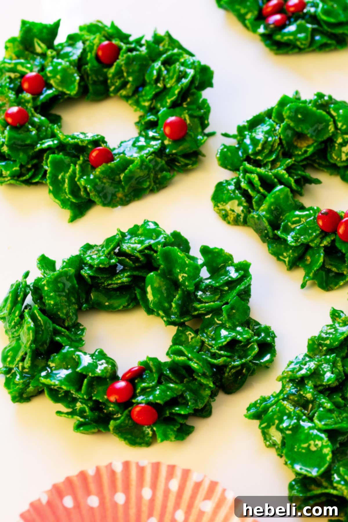 Close-up of several homemade Cornflake Christmas Wreaths, featuring their distinctive green color and red candy decorations, arranged on a white surface.