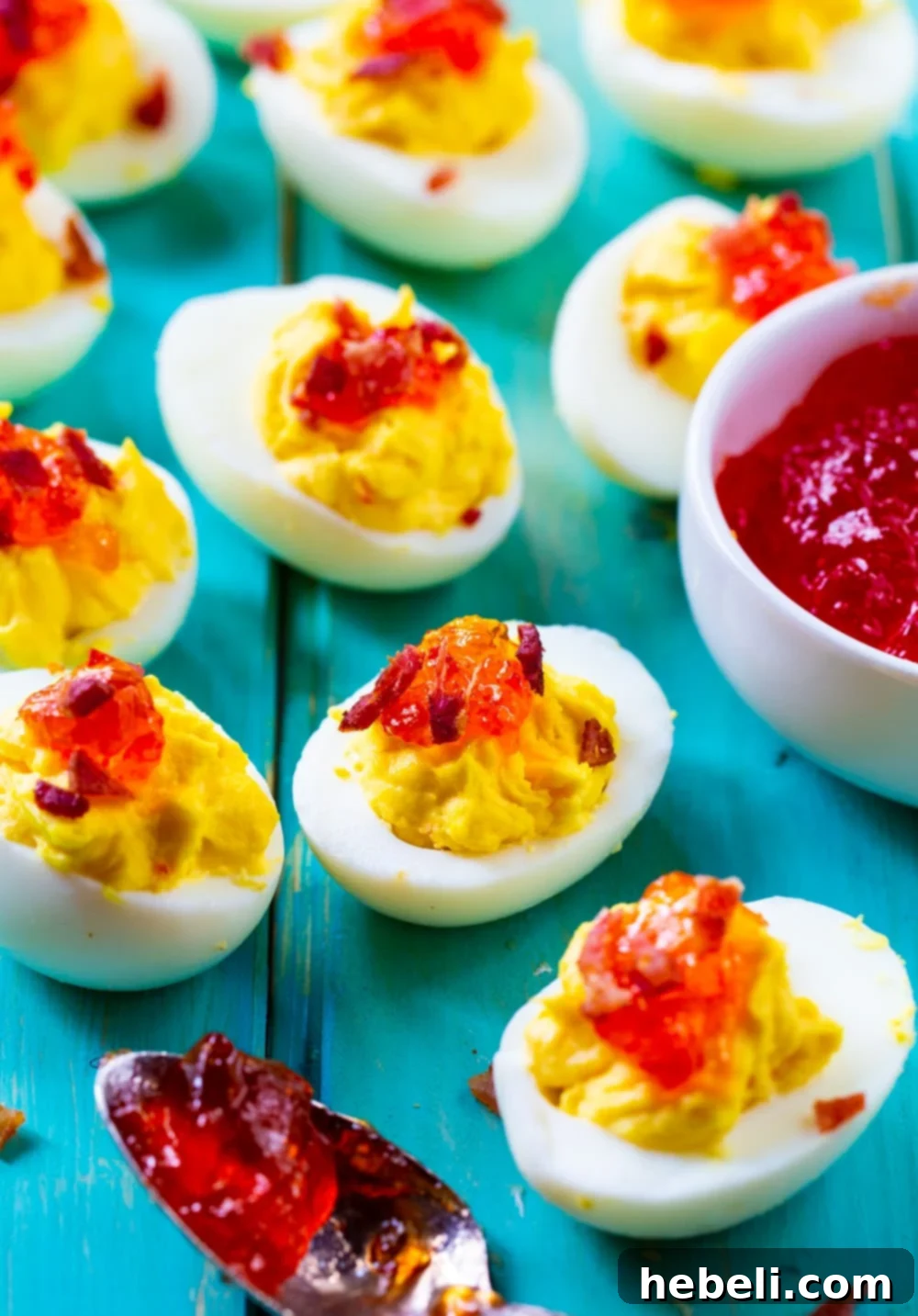 Smoky Bacon & Sweet Heat Deviled Eggs 3 Two halves of Red Pepper Jelly Deviled Eggs, each topped with a dollop of bright red jelly and crumbled bacon, set against a light blue backdrop.