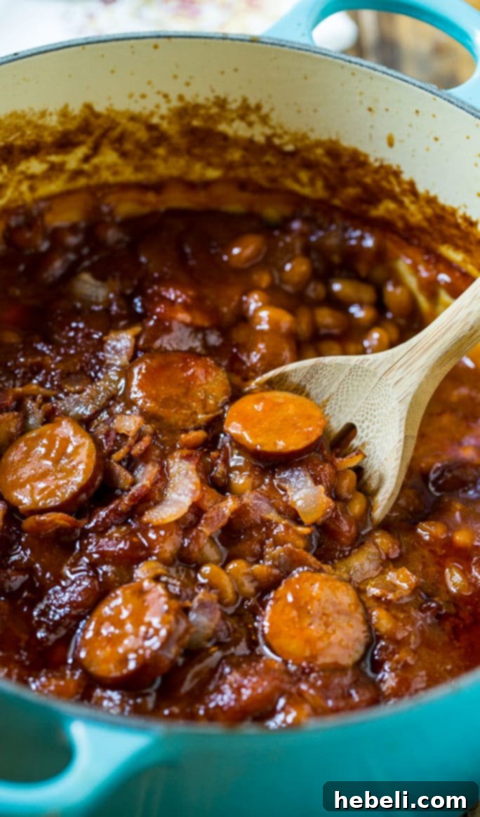 Close-up of hearty baked beans with visible pieces of smoked sausage and crispy bacon, ready to be served from a large casserole dish.