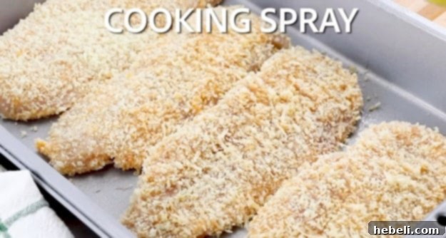 Hand spraying cooking oil over the breaded chicken breasts in the baking dish