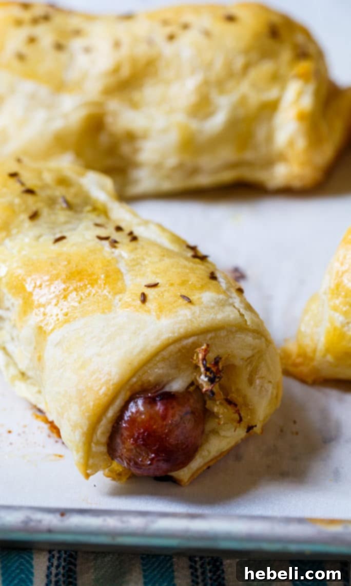 Four baked Brats in a Blanket on a baking sheet, golden brown and perfectly cooked, with caraway seeds visible on the pastry.