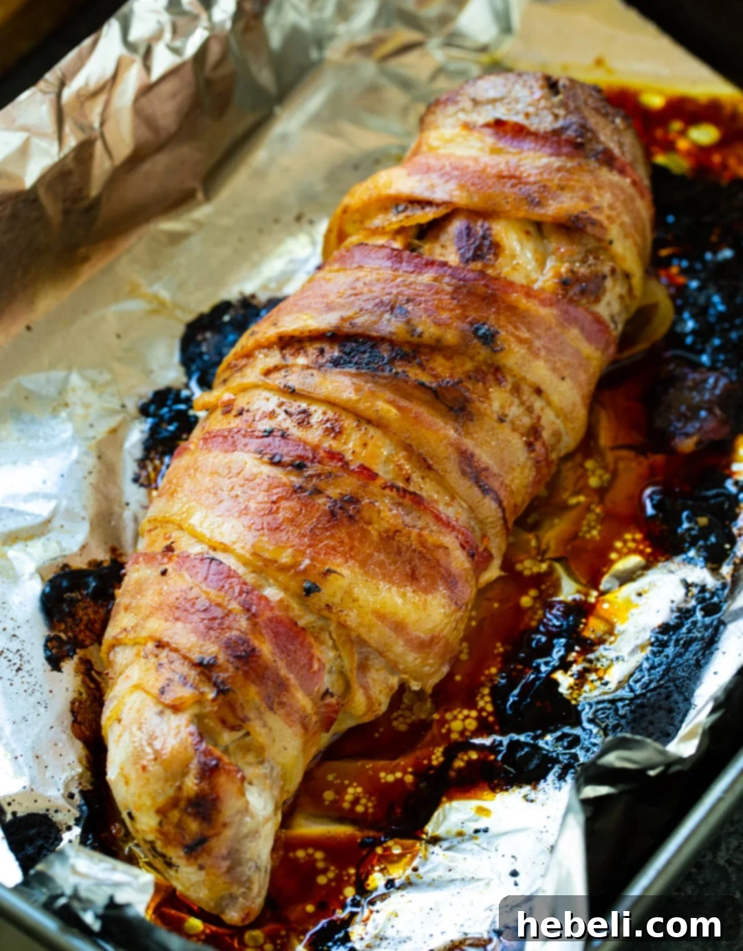 A golden brown, cooked pork tenderloin wrapped in crispy bacon, resting on a baking sheet after being removed from the oven.