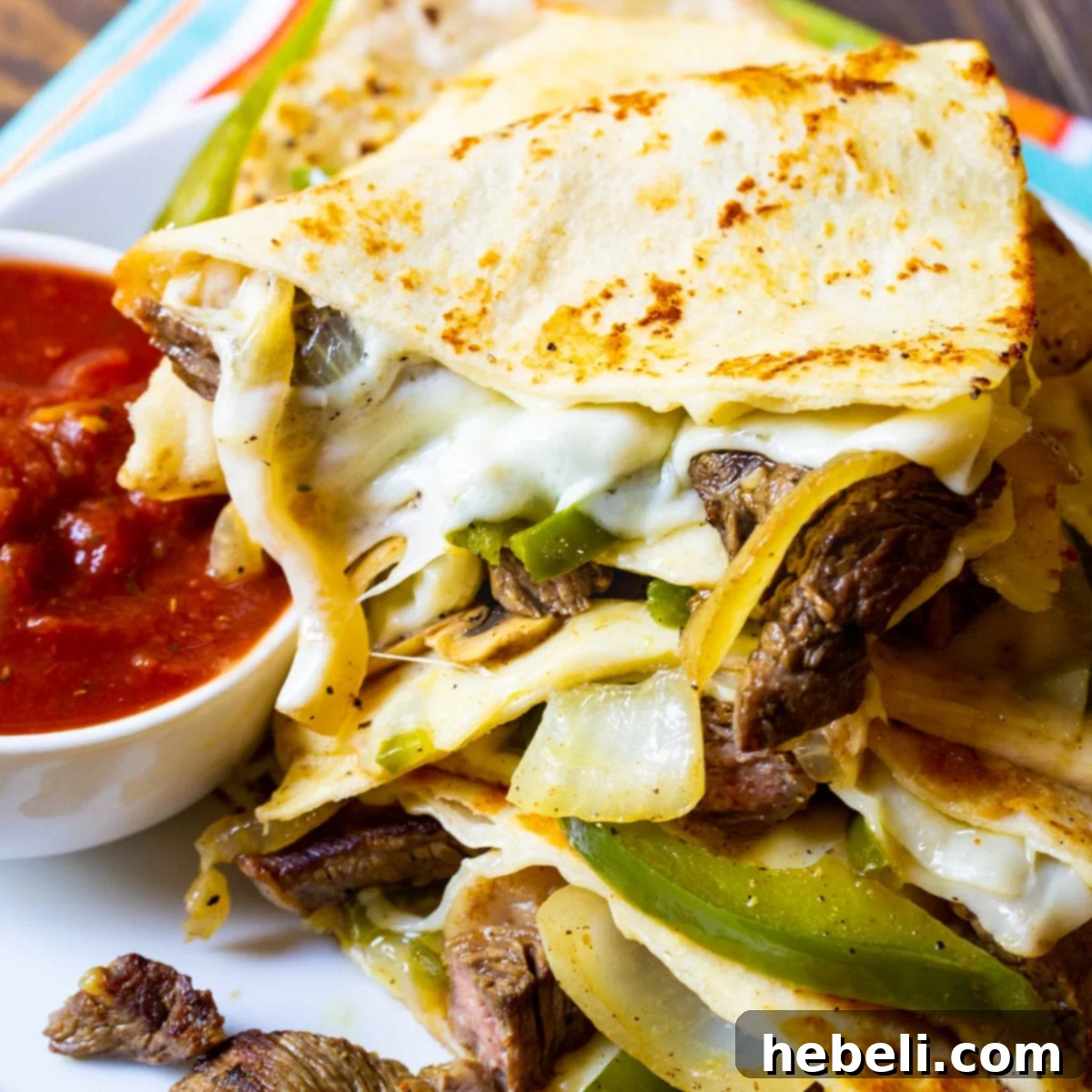 Triangles of Philly Cheese Steak Quesadilla piled up on a plate with a bowl of salsa, showcasing the melted cheese and filling.