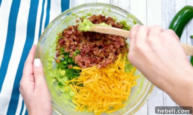 Folding diced jalapenos, shredded cheddar cheese, and crumbled bacon into the creamy guacamole base.