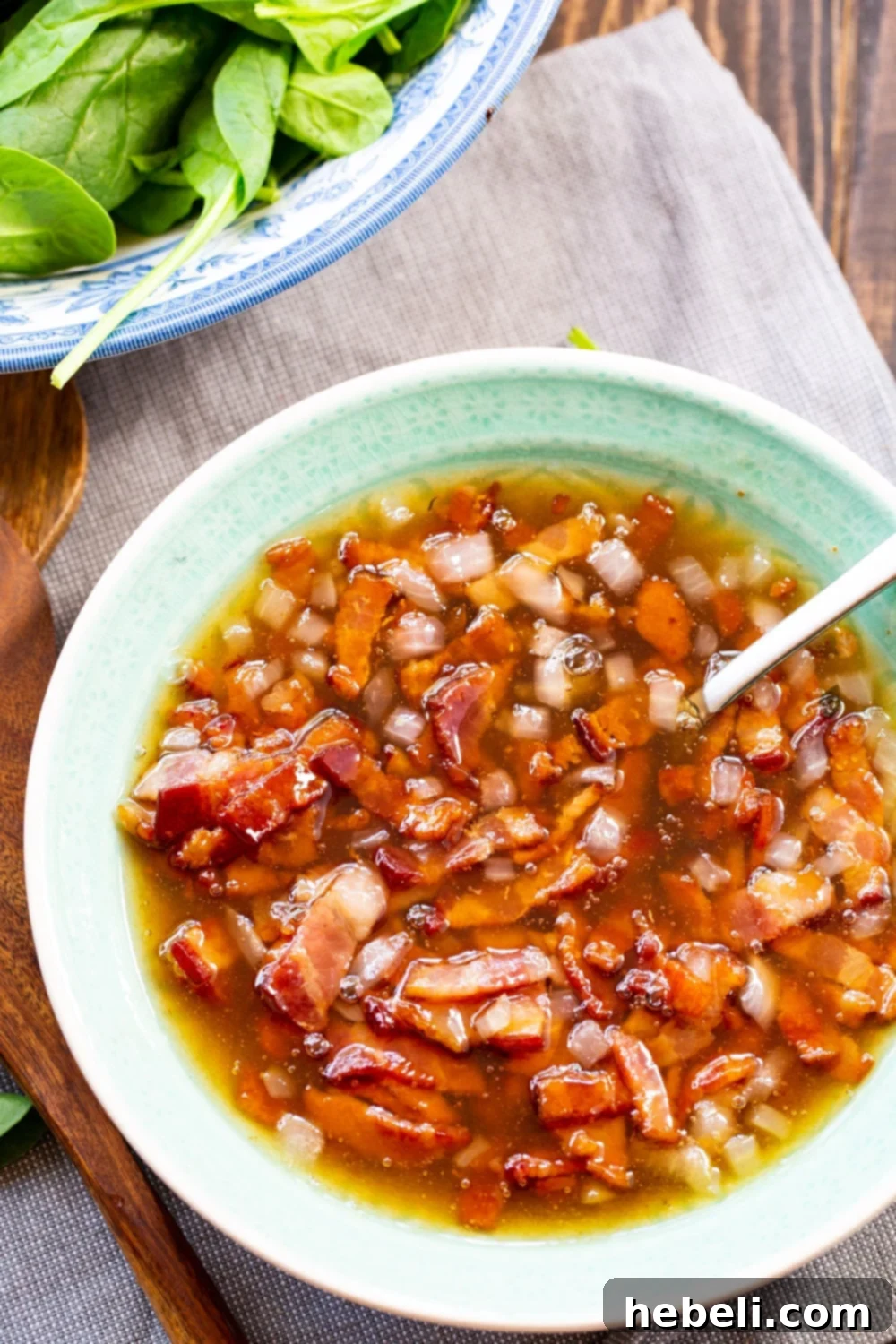 Close-up of Hot Bacon Dressing in a bowl, with a spoon resting in it, showcasing its rich texture and crispy bacon bits.