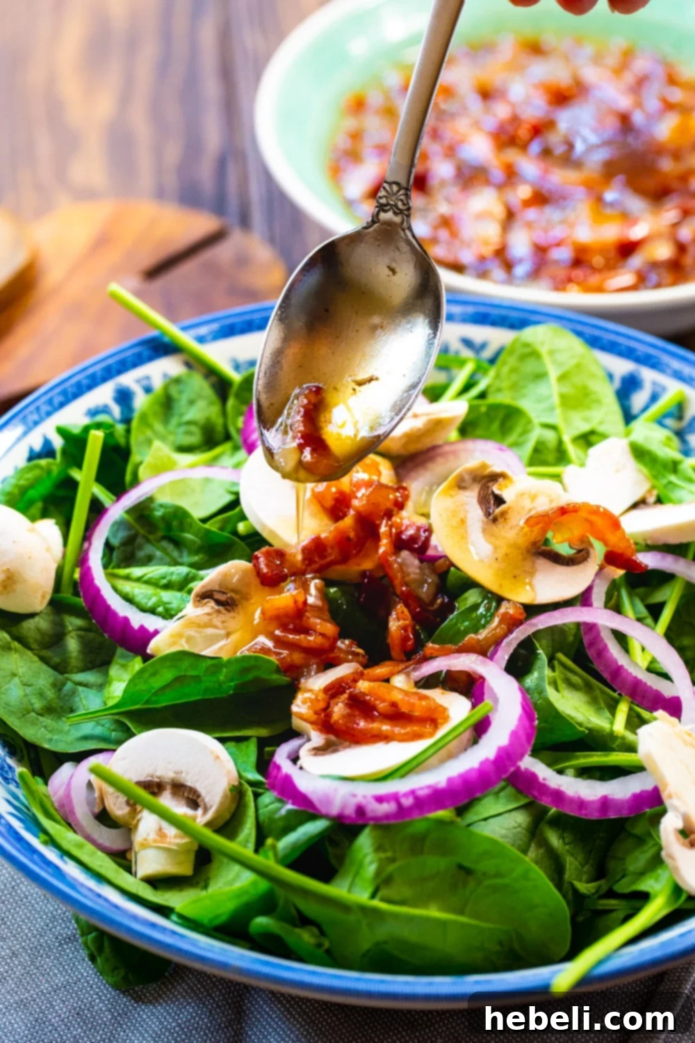 Hot Bacon Dressing being spooned generously over a fresh salad of spinach, mushrooms, and red onion.