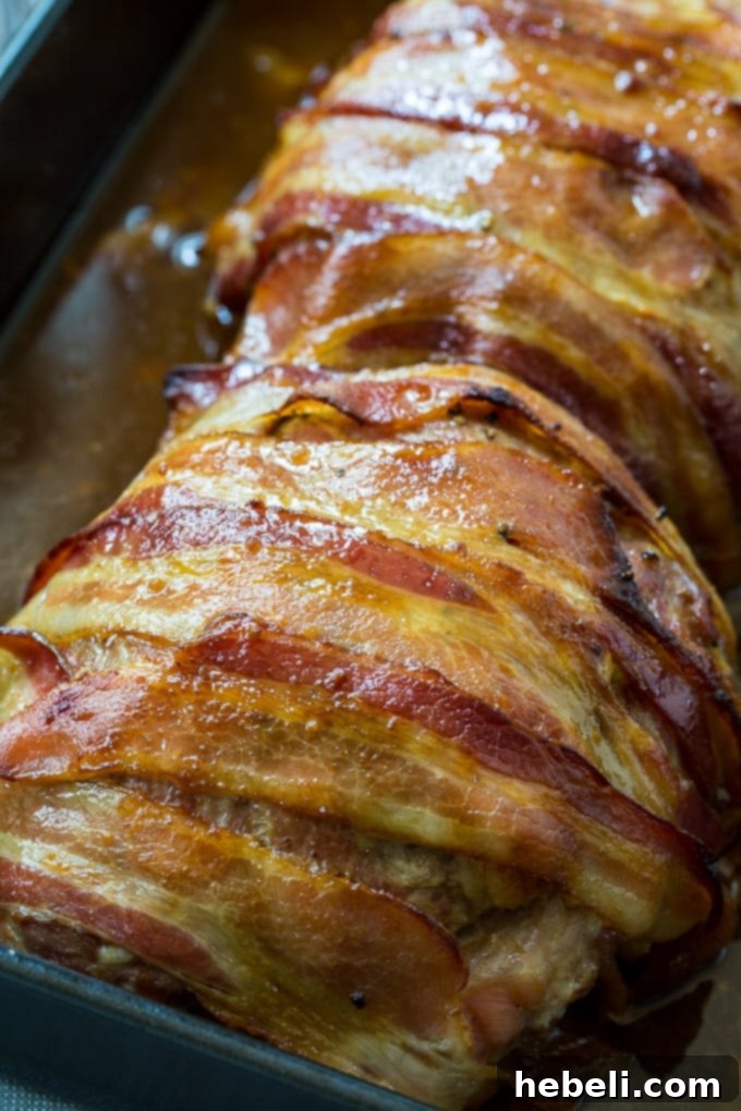 Raw pork loin coated in maple glaze, being wrapped with bacon slices.