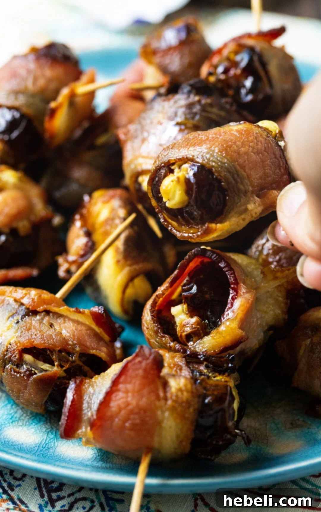 Sweet and Savory Bacon Wrapped Stuffed Dates 5 A hand reaching for a perfectly cooked Bacon Wrapped Stuffed Date from a serving platter.
