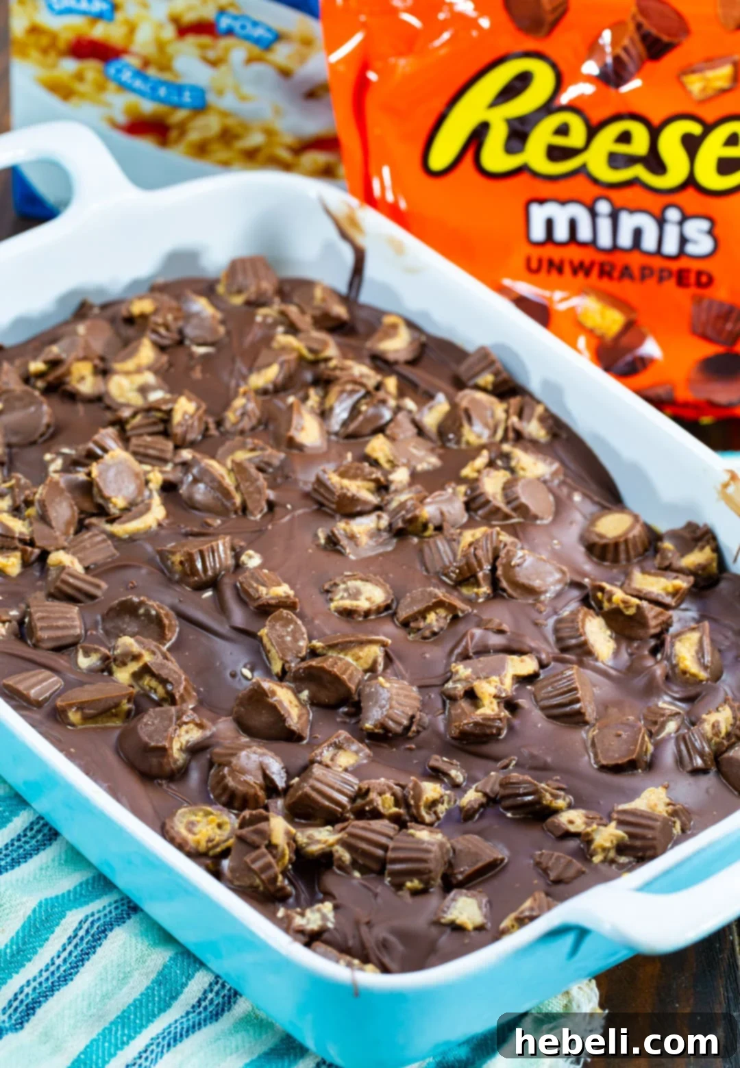 No-Bake Chocolate Peanut Butter Crispy Squares 3 A close-up of freshly made Chocolate Peanut Butter Rice Krispie Treats in a baking pan, showing the even layer of chocolate and peanut butter topping with visible mini peanut butter cups, ready to be chilled and sliced.