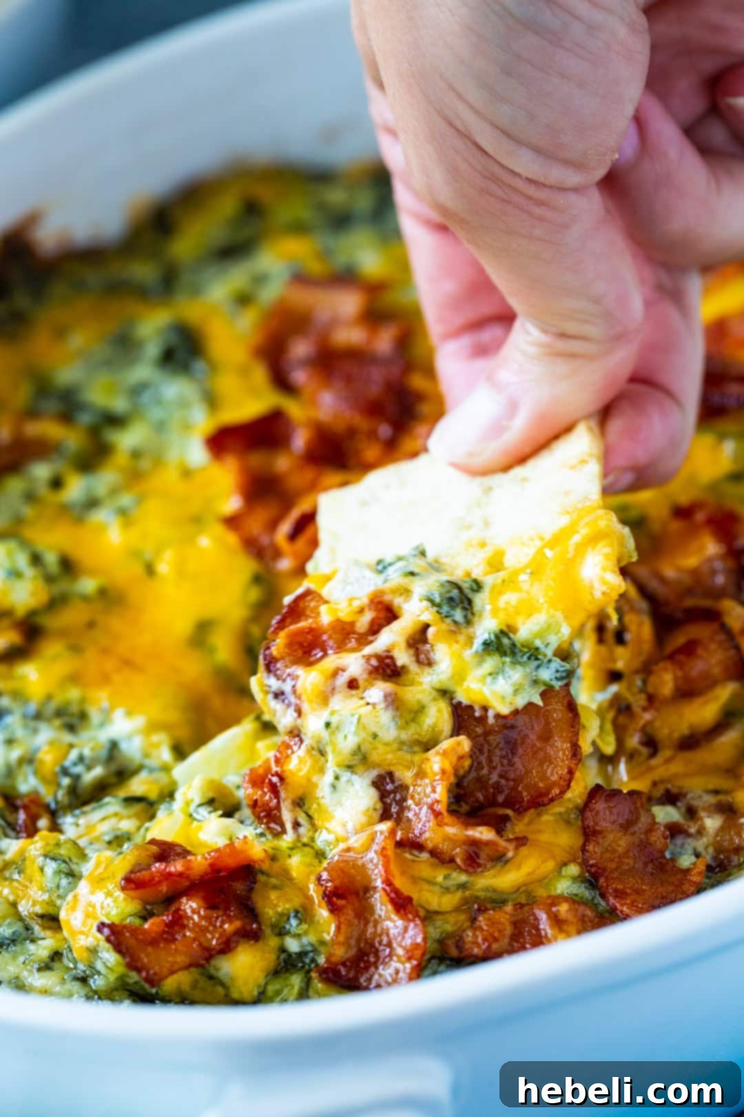 Hand reaching for a scoop of Warm Turnip Green Dip with a crispy pita chip, showcasing the dip's rich texture.