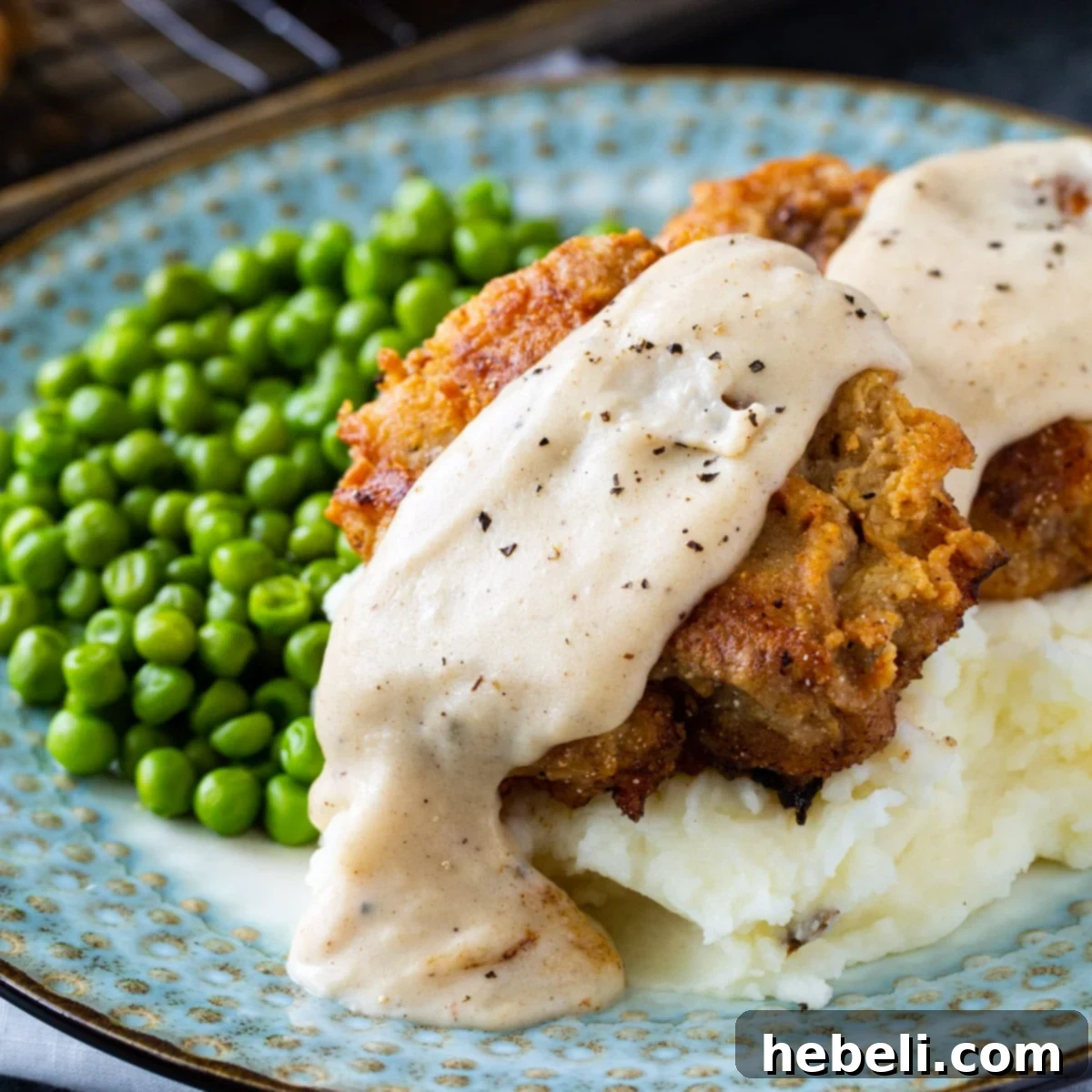 Crispy Chicken Fried Hamburger patties with rich white gravy served over fluffy mashed potatoes, accompanied by green peas.