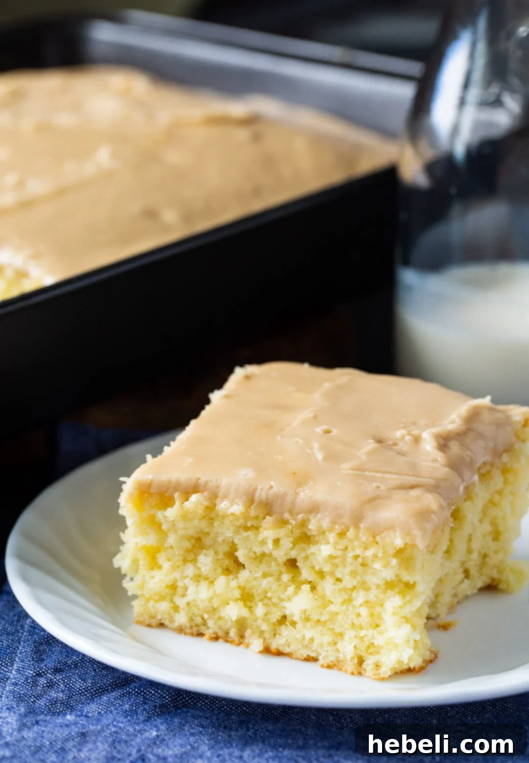 A close-up of a generous slice of Buttermilk Sheet Cake with creamy caramel icing, ready to be enjoyed, with the rest of the cake in the background.