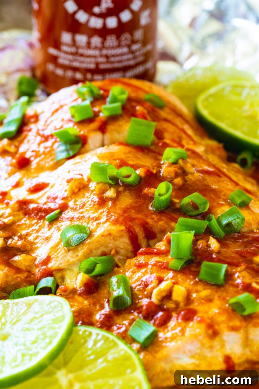 Close-up of baked Firecracker Salmon with lime slices and a bottle of Sriracha, highlighting the vibrant colors and delicious glaze.