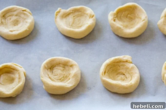 Crescent roll dough slices being shaped by hand into shallow bowls on a baking sheet, preparing them to hold the filling.