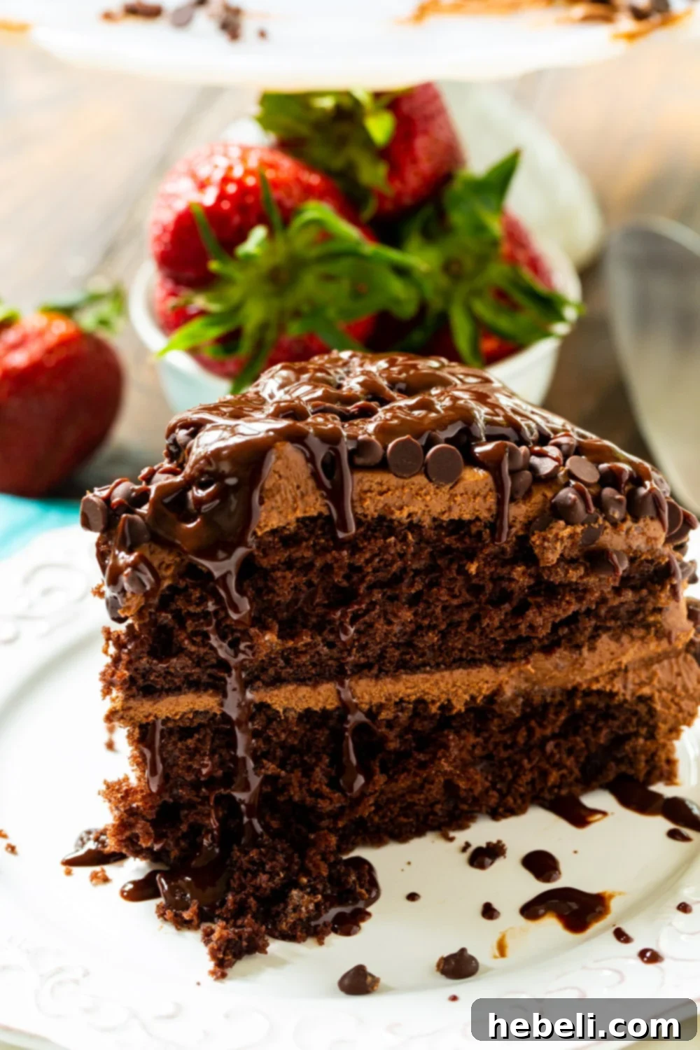 A generously sized slice of Death by Chocolate Cake presented with fresh, vibrant strawberries, highlighting its rich, multi-layered chocolate construction and creamy frosting.