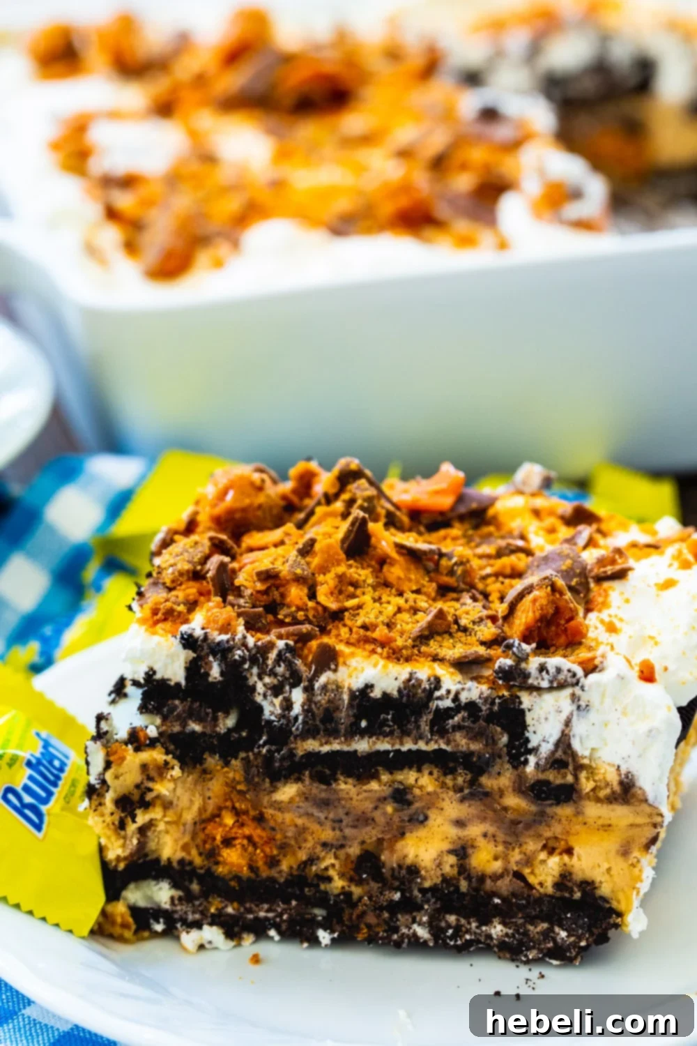 Ultimate No-Bake Butterfinger Oreo Delight 3 A close-up shot of a slice of the icebox cake on a plate, with the remainder of the cake in a baking dish in the blurred background, highlighting its irresistible texture.