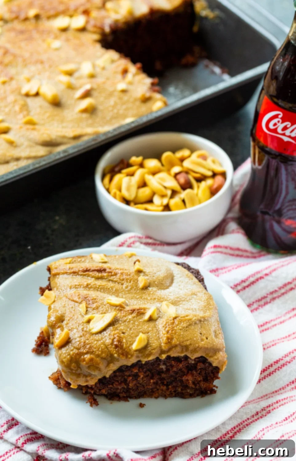 A beautifully presented slice of Coca-Cola Cake, showing the distinct layers of moist chocolate cake and the caramelized, broiled peanut frosting, with the rest of the sheet cake visible in the background.