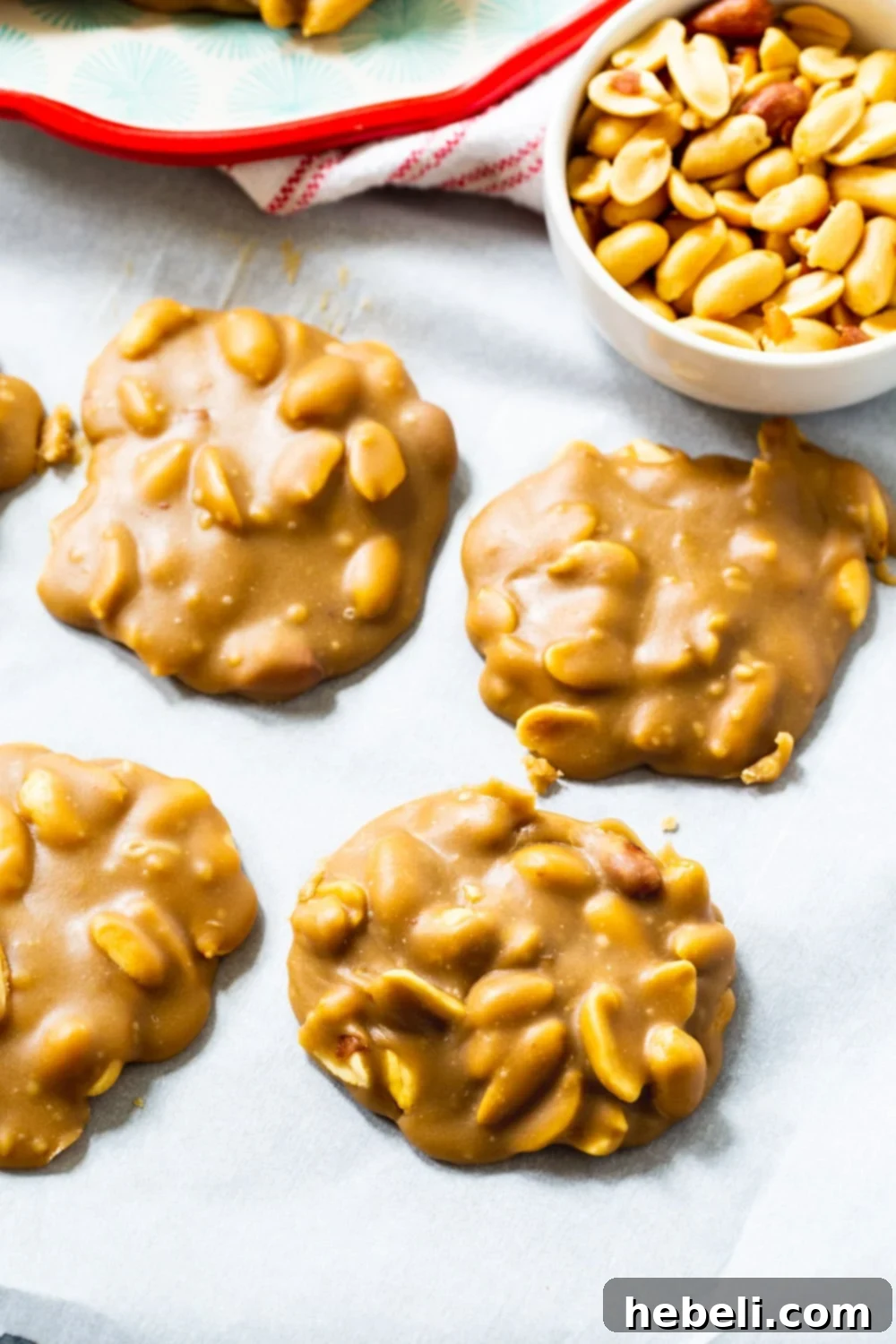 Freshly dropped Peanut Pralines cooling on parchment paper, with a bowl of roasted peanuts in the background.