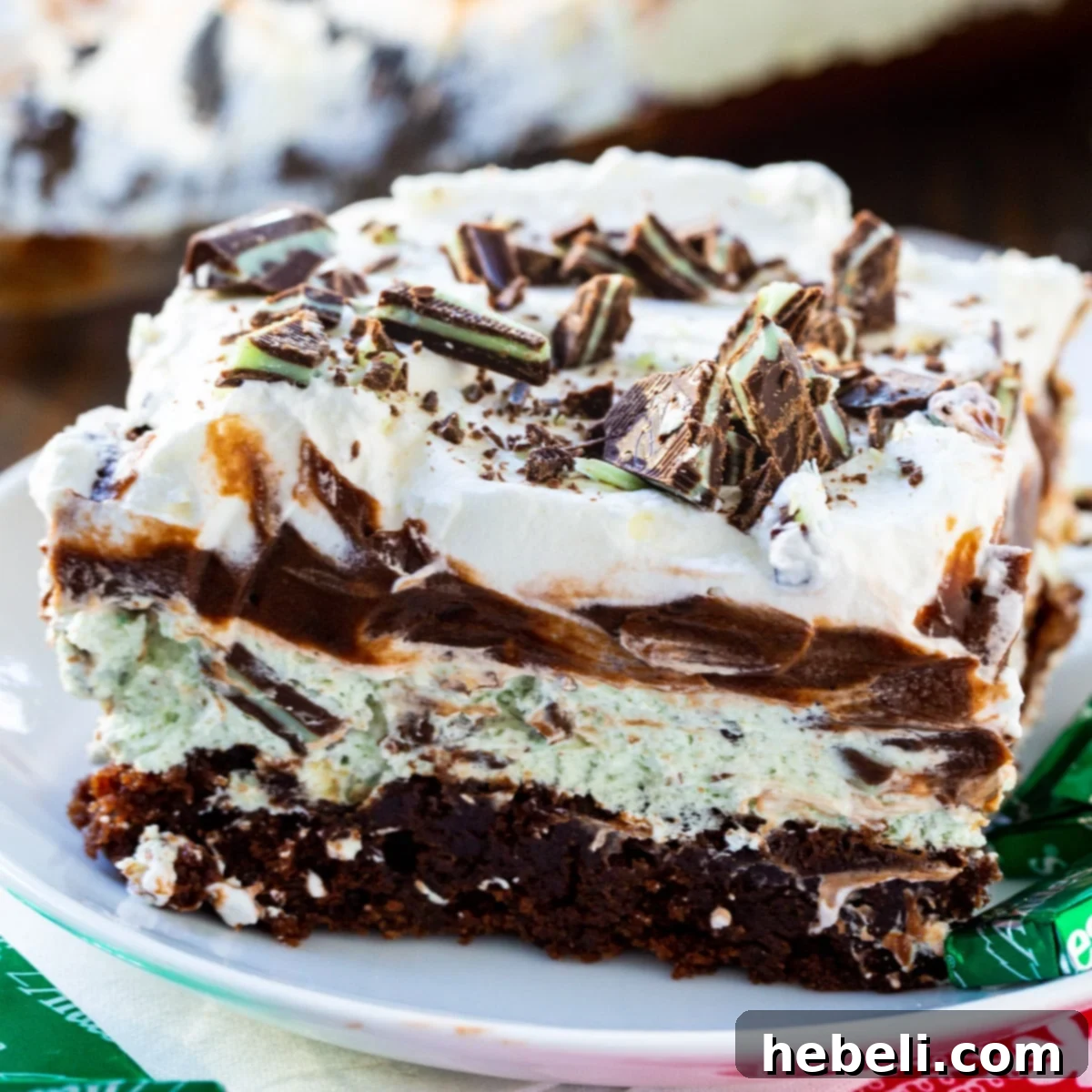 A delectable slice of Andes Mint Lush on a pristine white plate, showcasing its four distinct layers of fudgy brownie, mint cream cheese, rich chocolate pudding, and fluffy whipped topping, garnished with crushed Andes Mints.