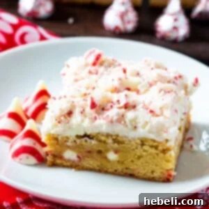 A frosted blondie on a small white plate, garnished with peppermint pieces, with more unwrapped Hershey's Peppermint Kisses beside it.