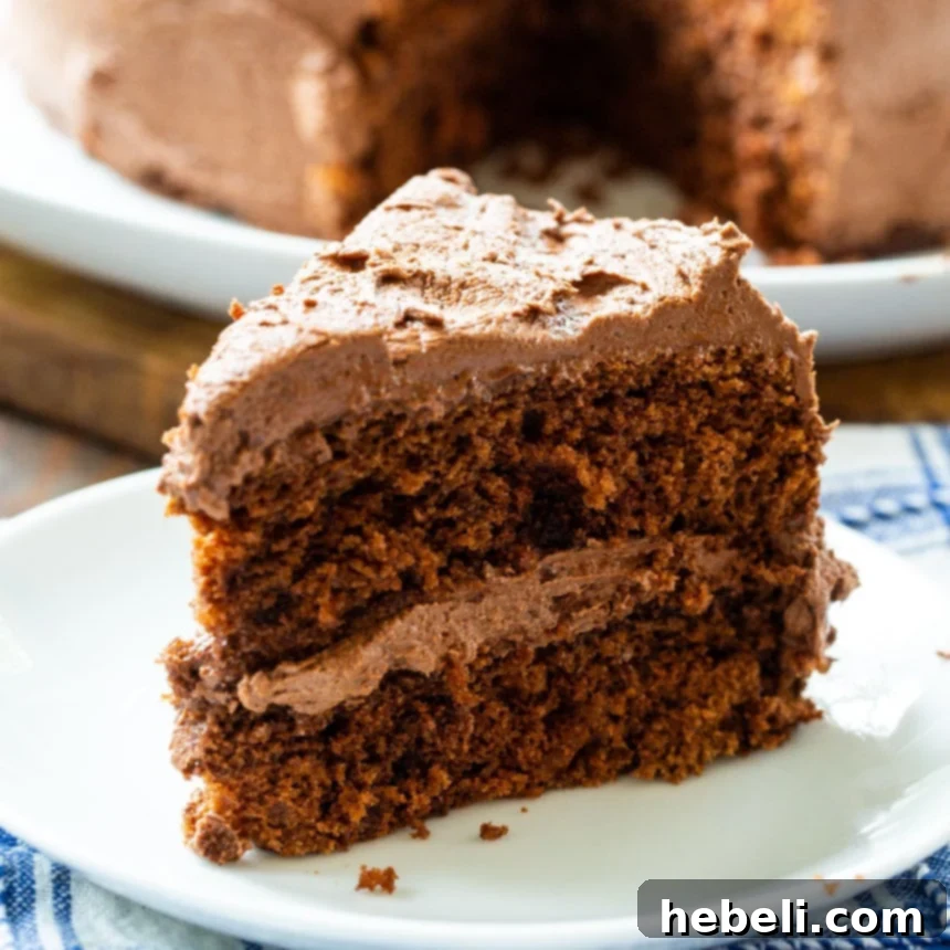 A delectable slice of Classic Chocolate Layer Cake on a small white plate, showcasing its moist, dark layers and thick, smooth chocolate buttercream frosting.