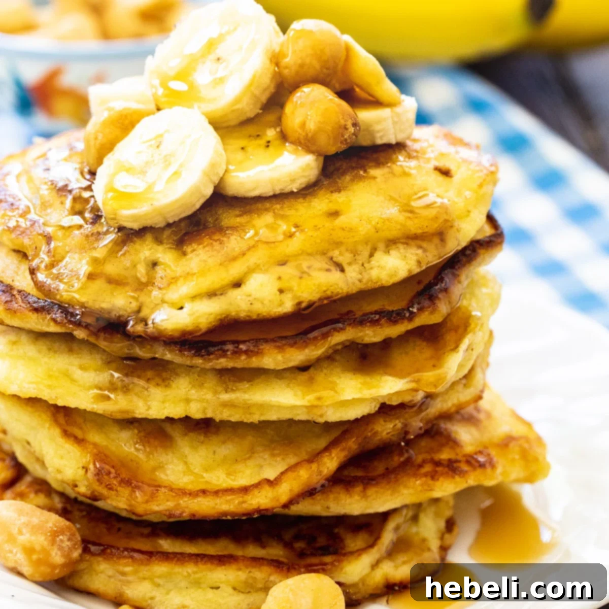 Stack of golden-brown Banana Macadamia Pancakes with syrup, garnished with fresh banana slices and a sprinkle of macadamia nuts.