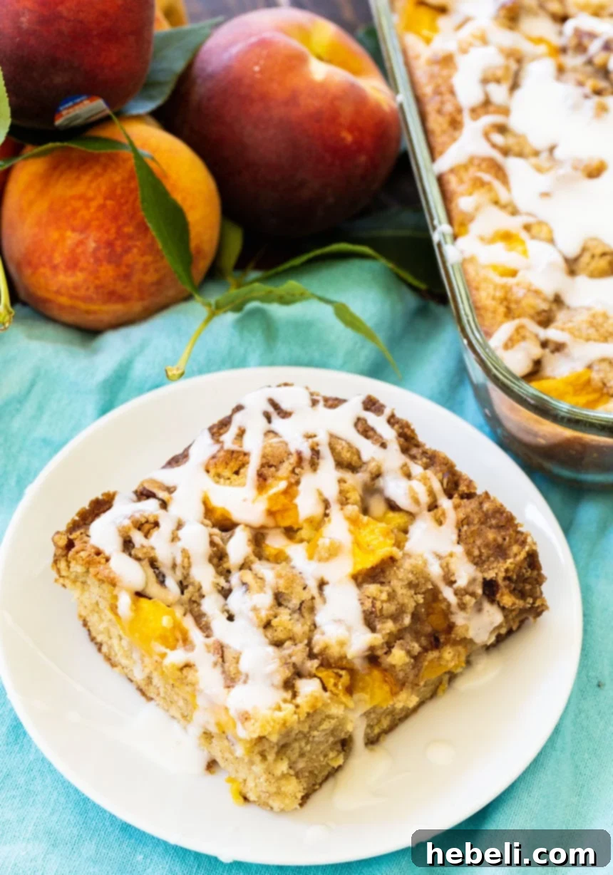 Peach Crumble Coffee Cake 3 A close-up shot of a slice of Peach Crisp Coffee Cake on a white plate, with whole fresh peaches blurred in the background, highlighting the golden brown streusel and delicate glaze.
