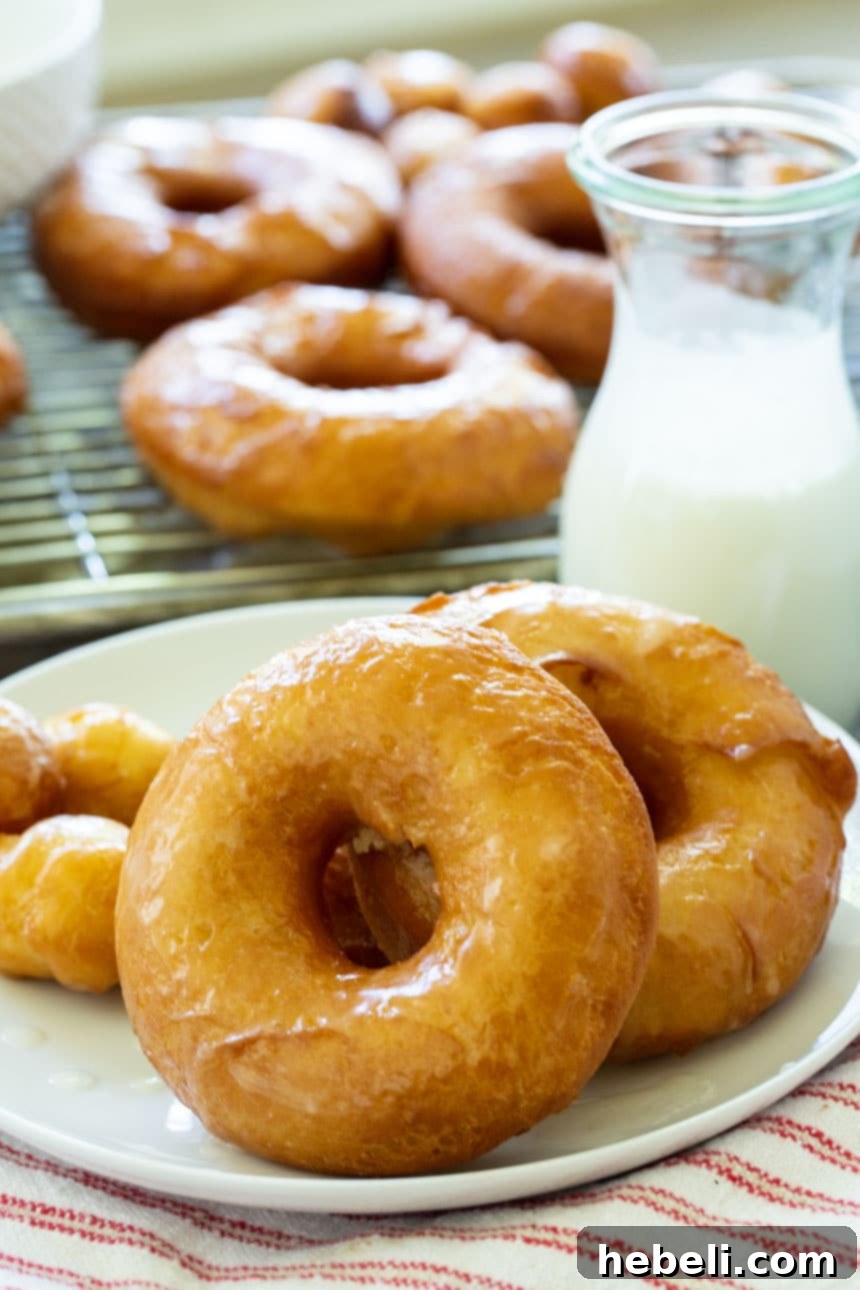 Homemade Krispy Kreme Style Doughnuts 4 Close-up of perfectly glazed Copycat Krispy Kreme Doughnuts on a plate, with more in the background, showcasing their irresistible shine and fluffy interior.