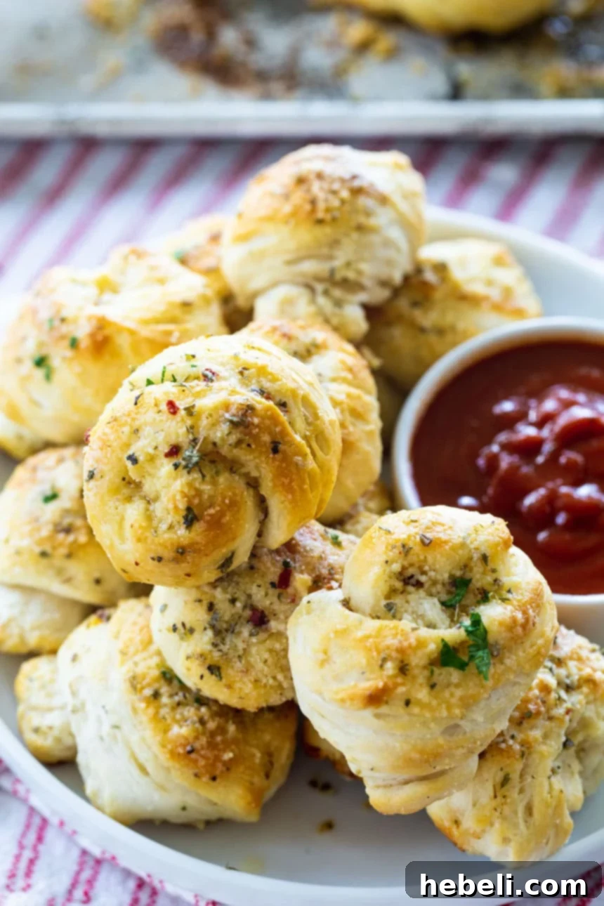 A stack of golden easy garlic knots on a white plate, with a small bowl of pizza sauce for dipping.