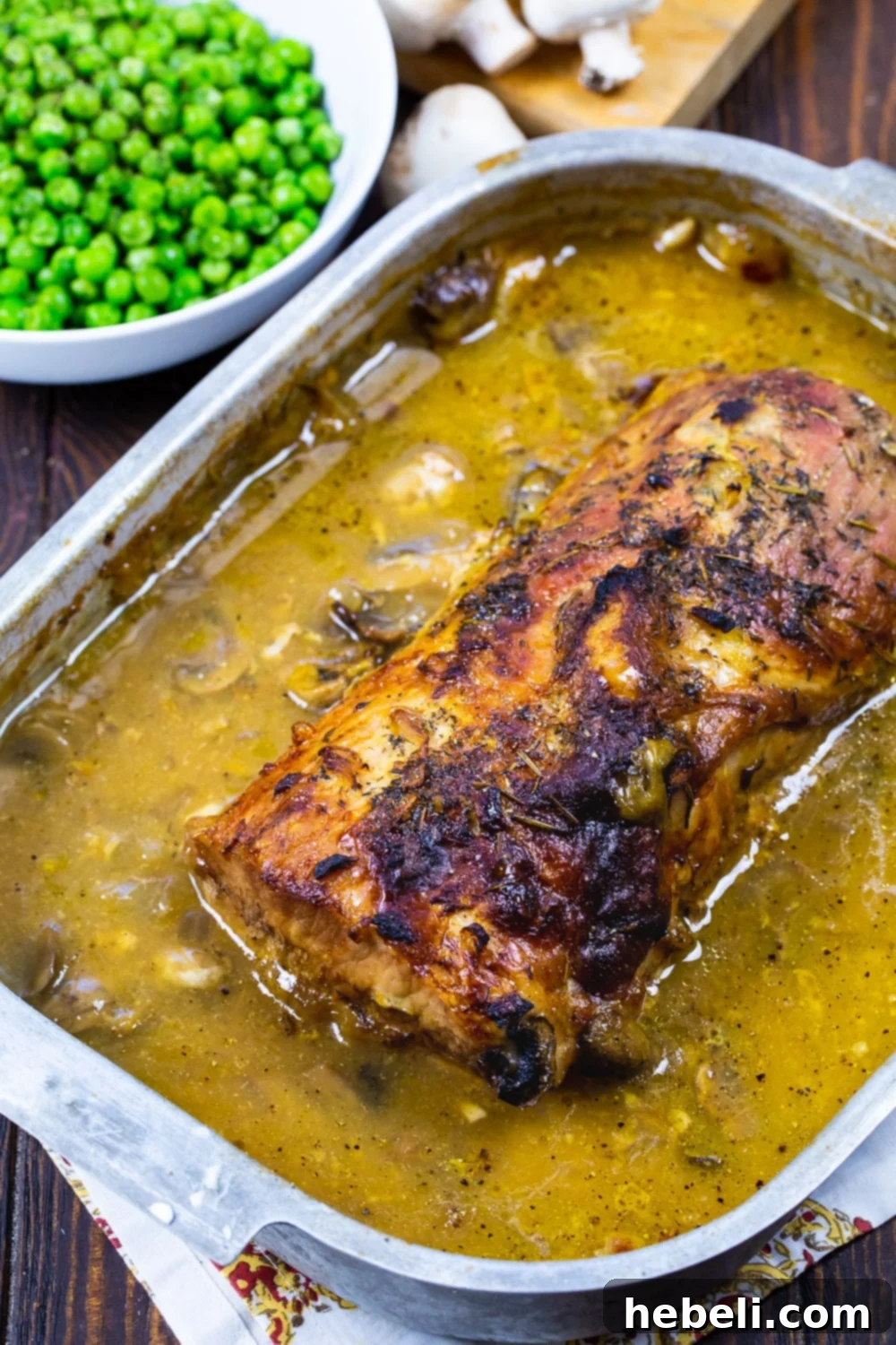Close-up of an uncooked pork loin in a heavy roasting pan, seasoned with herbs and spices, surrounded by raw mushrooms and onions, ready for the oven.