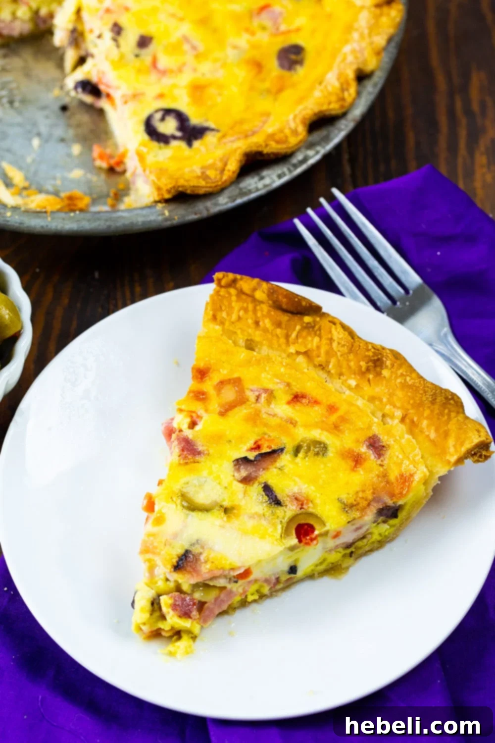 Savory Muffaletta Quiche 3 Muffaletta Quiche baking in a pie pan, with a finished slice on a plate beside it, showing the rich filling.