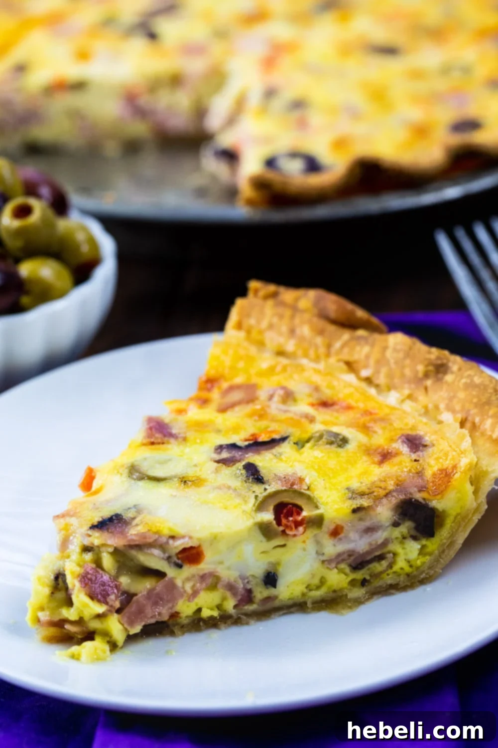 Savory Muffaletta Quiche 4 A beautifully presented slice of Muffaletta Quiche, rich with olives, ham, salami, and mortadella on a white plate.