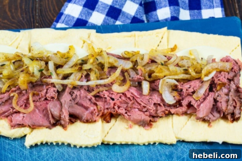 Flavorful French Dip Roll-Ups 4 Crescent roll dough rectangles laid out, topped with slices of Provolone cheese, deli roast beef, and a generous layer of caramelized onions, ready to be rolled.