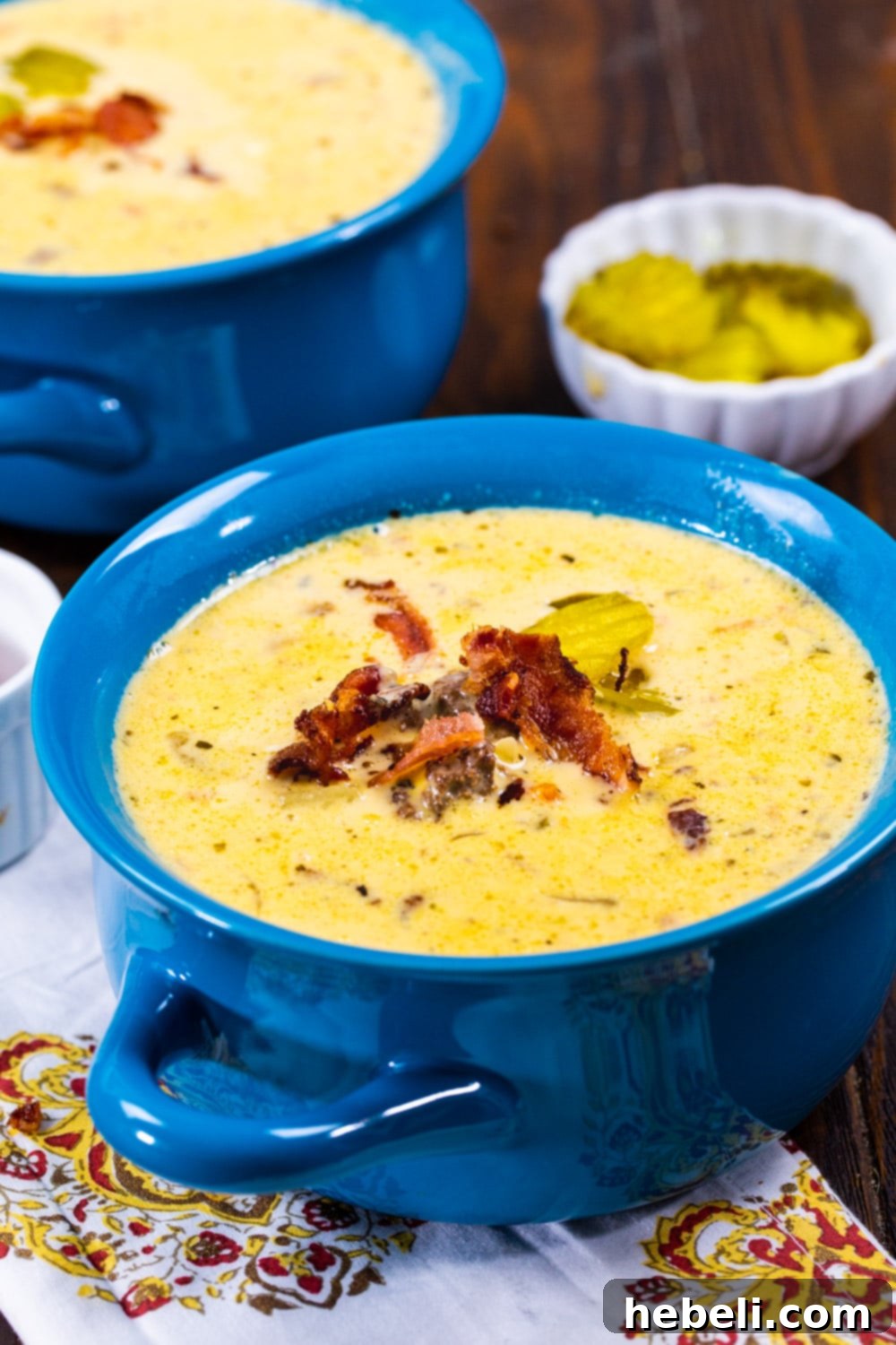 Loaded Slow Cooker Bacon Cheeseburger Soup 4 A vibrant blue bowl filled to the brim with creamy Slow Cooker Bacon Cheeseburger Soup, garnished with crumbled bacon and a few pickle slices.