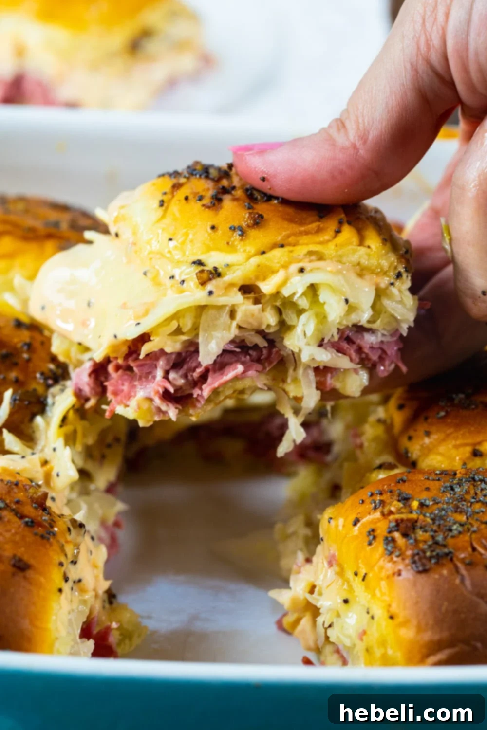 A hand reaches for a Baked Reuben Slider, showcasing the melted cheese and juicy corned beef on a sweet Hawaiian roll.