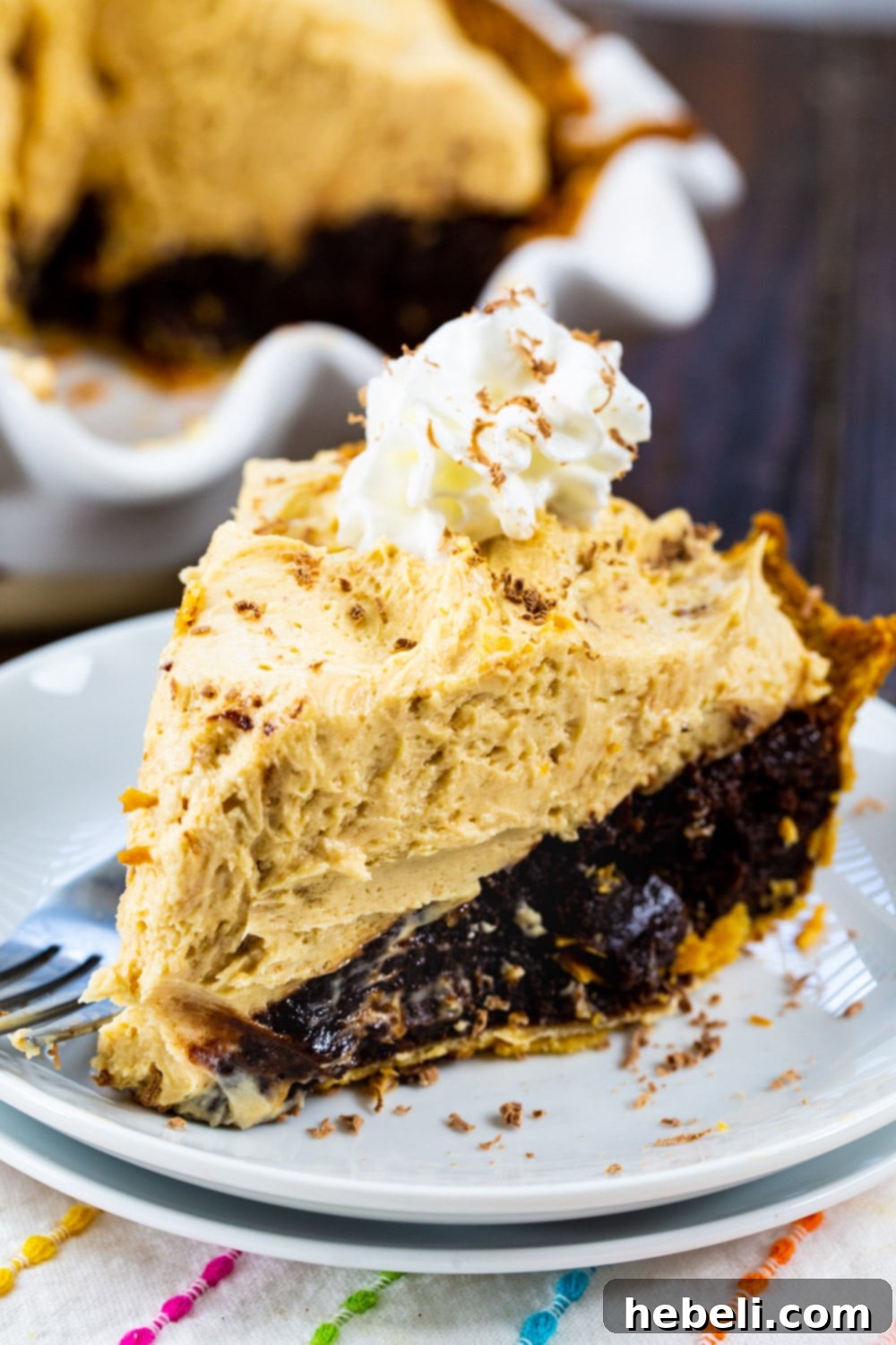 A close-up of a single slice of Peanut Butter Mousse Brownie Pie on a white plate, with the rest of the pie blurred in the background, highlighting the individual serving.