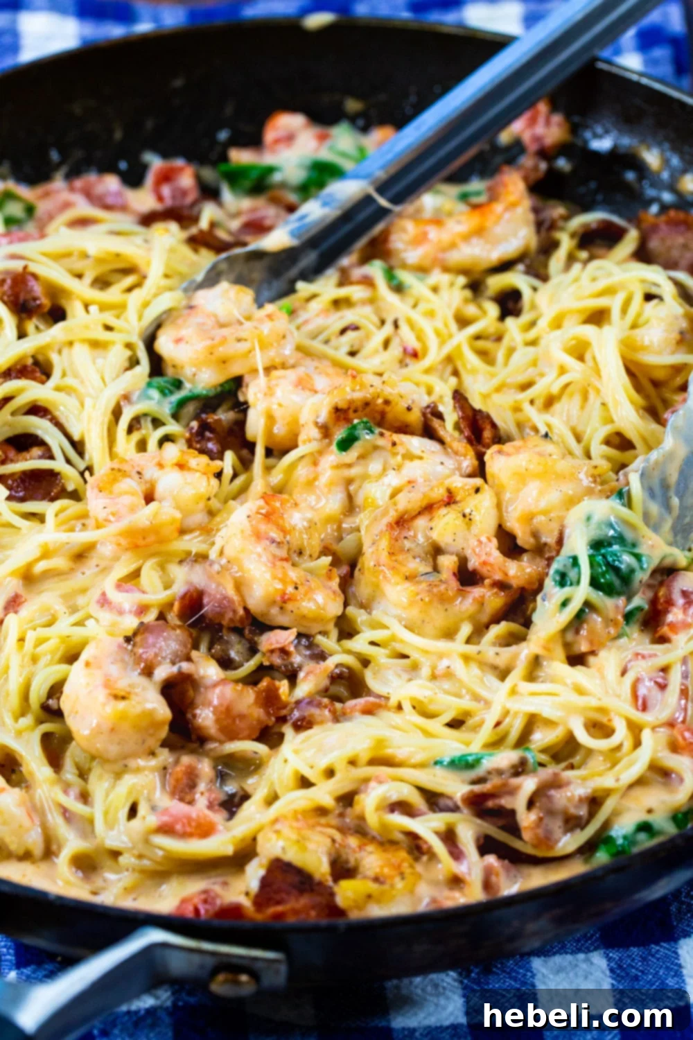 Tongs gently scooping a generous portion of Creamy Angel Hair Pasta with Bacon and Shrimp from a skillet.