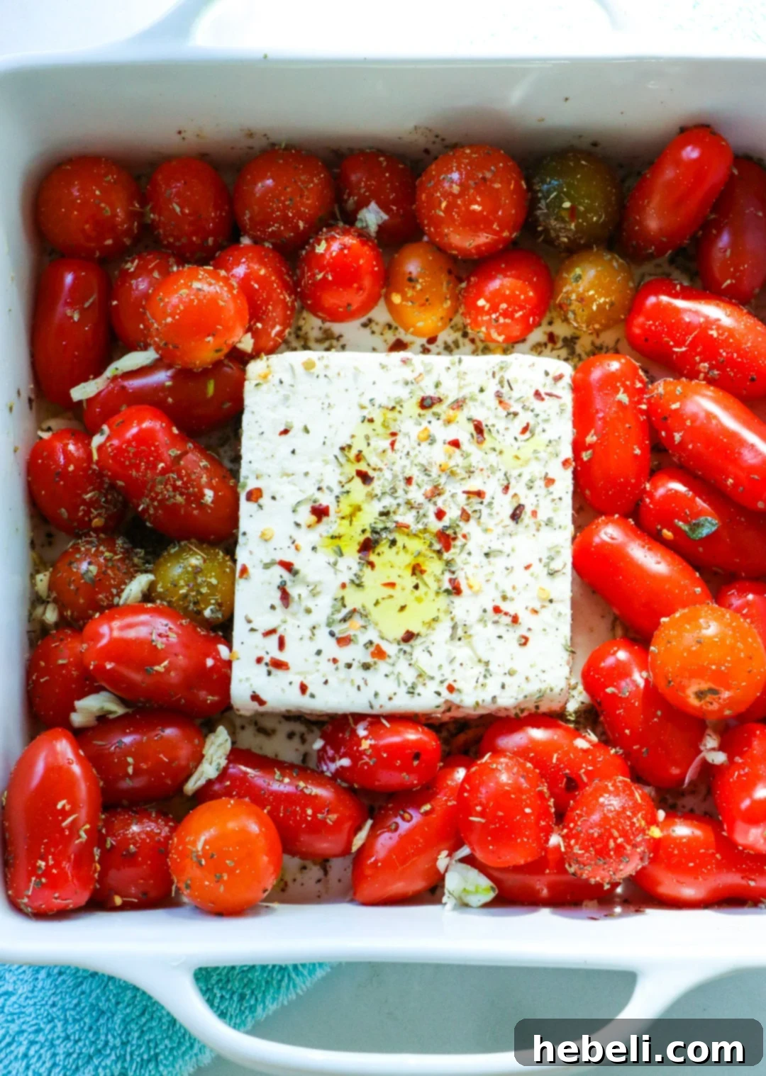 Close-up of cherry tomatoes and a solid block of feta cheese, seasoned and drizzled with olive oil, arranged in a square baking dish, prior to baking.
